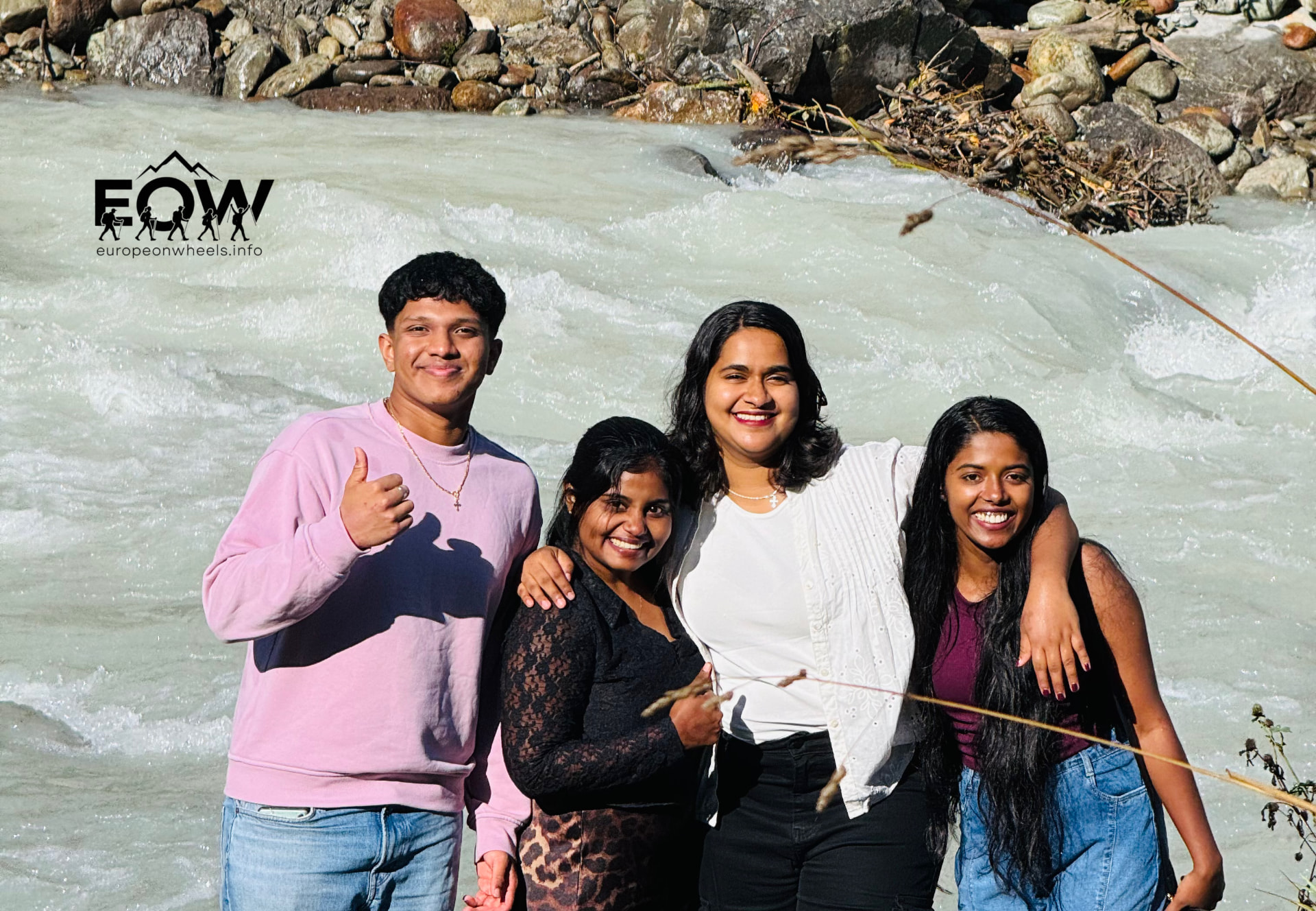 Four travellers smiling together by the glacier-blue river in the Swiss valley