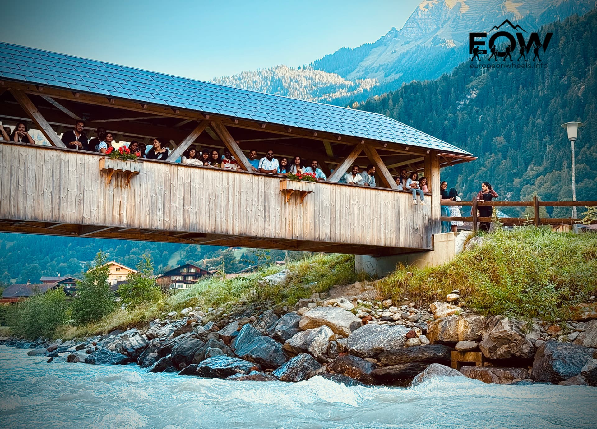 Travellers on the flower-lined covered bridge with alpine peaks behind, Switzerland